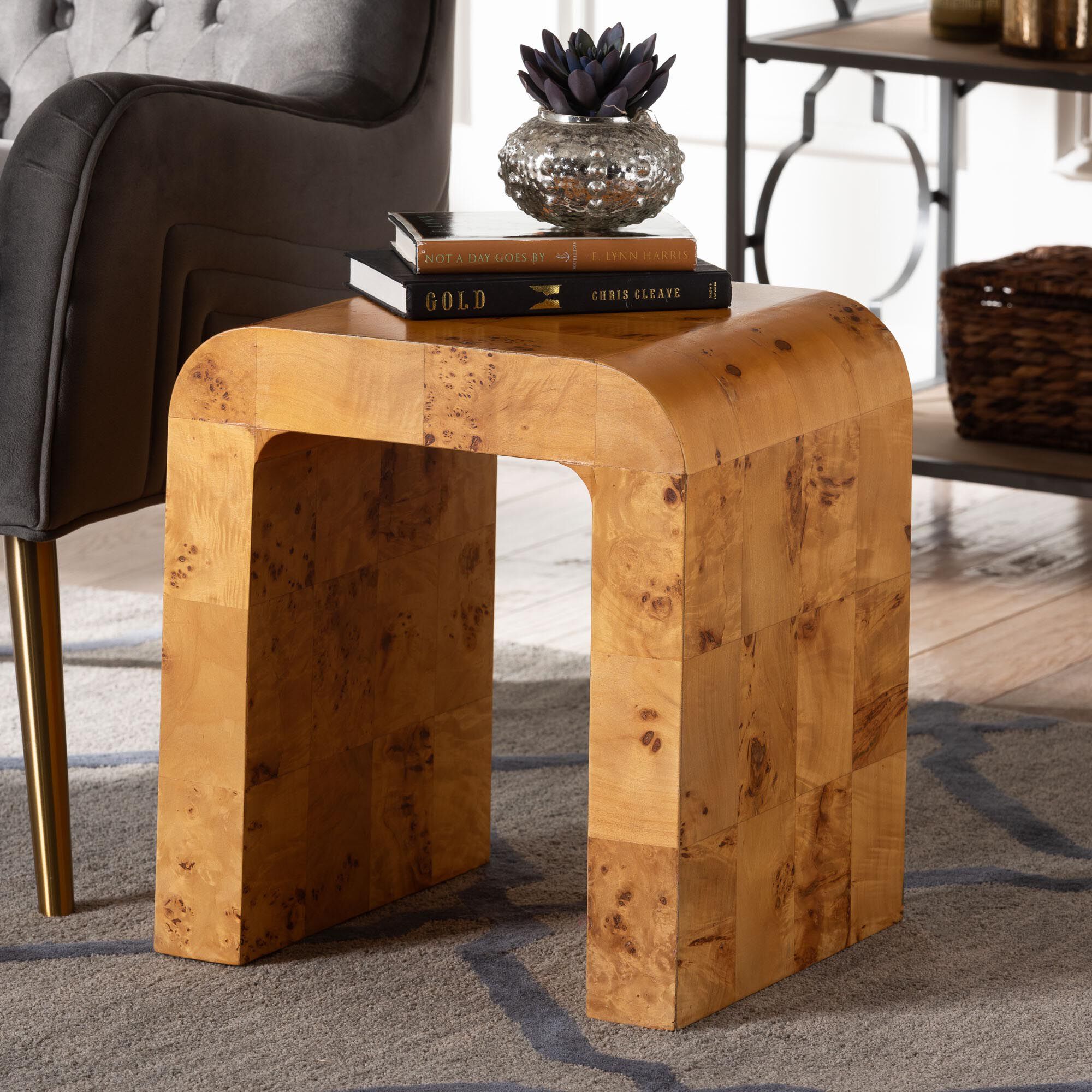 The burl wood end table in a living room setting with a chair and shelf in the background.